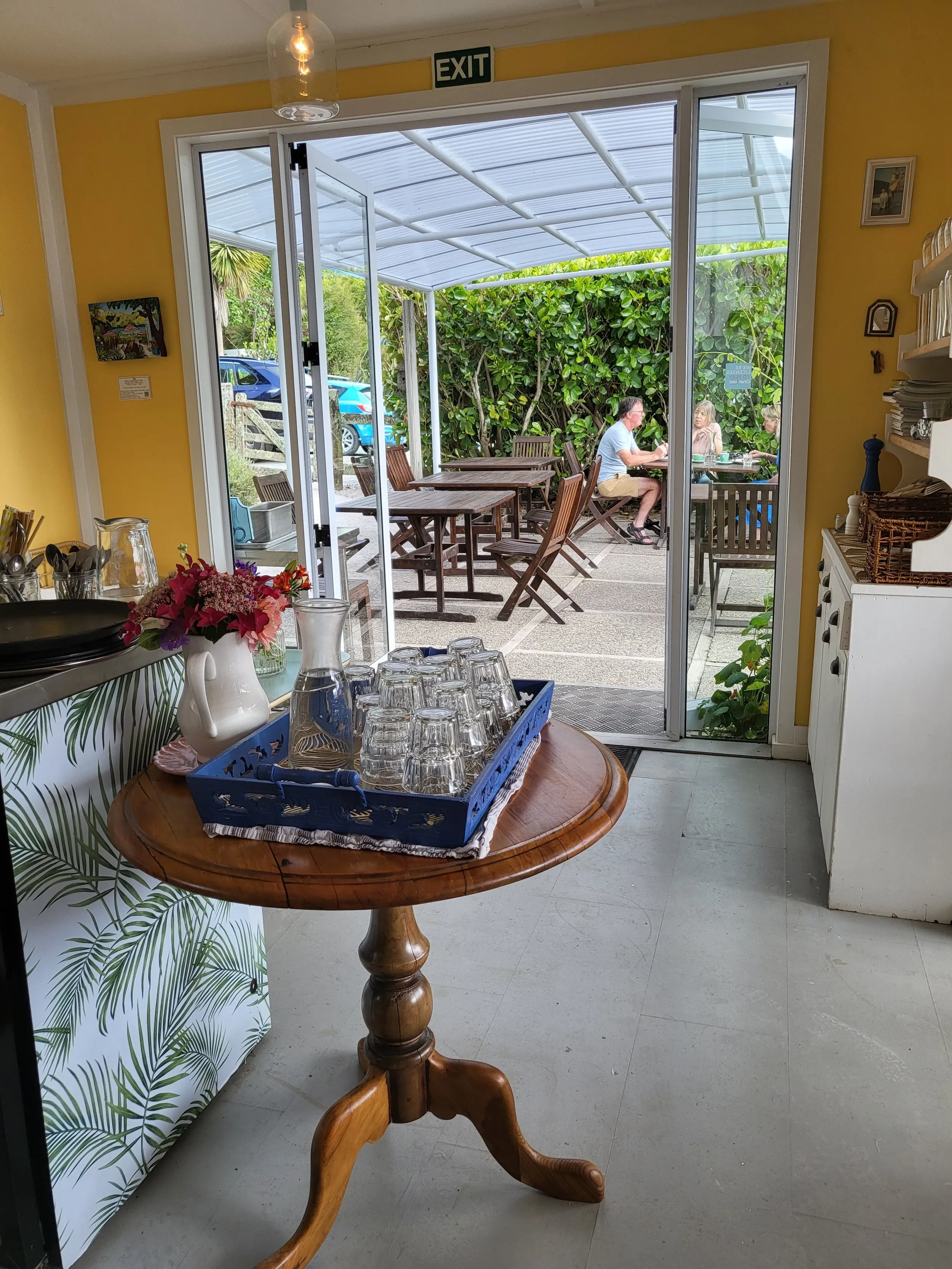 Looking from inside past a table of glasses and flowers, through glass doors to people eating on the covered deck