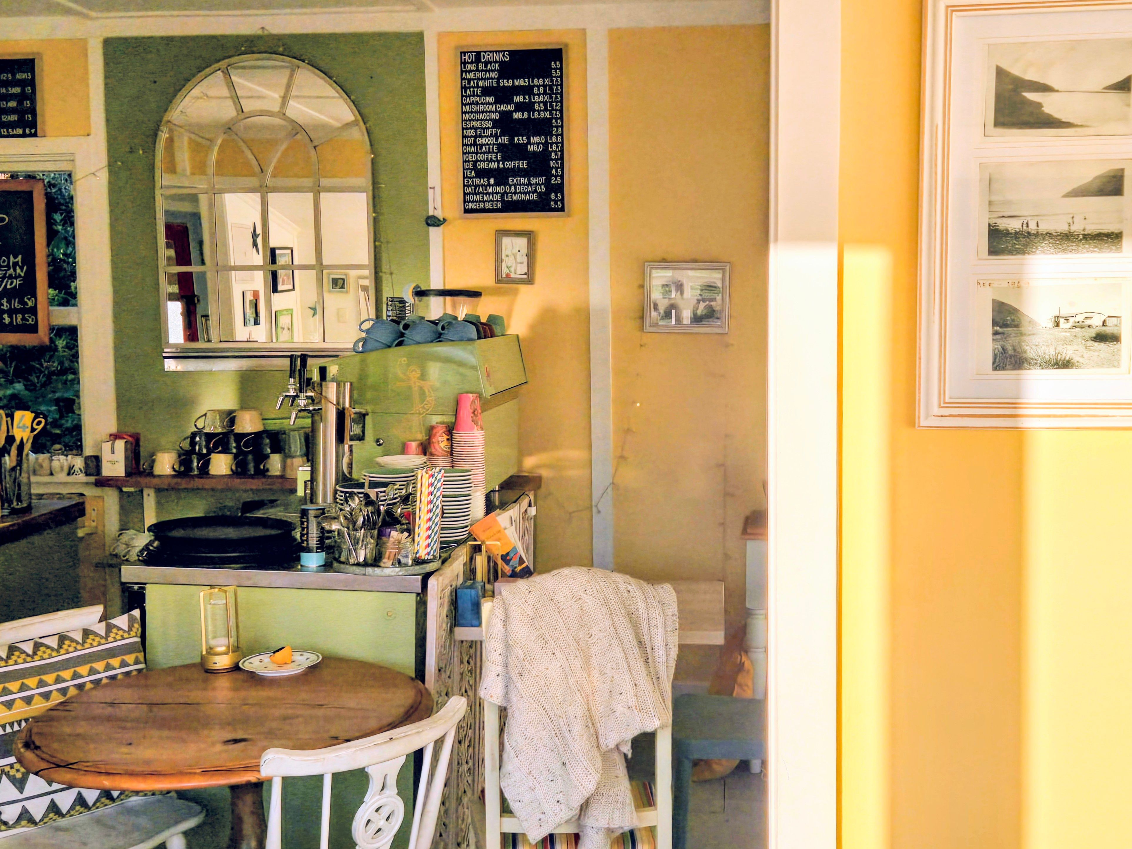 Inside the café in the afternoon sun: the green coffee machine under the hot drinks board, a round wooden table, and old Cable Bay photos on the yellow wall