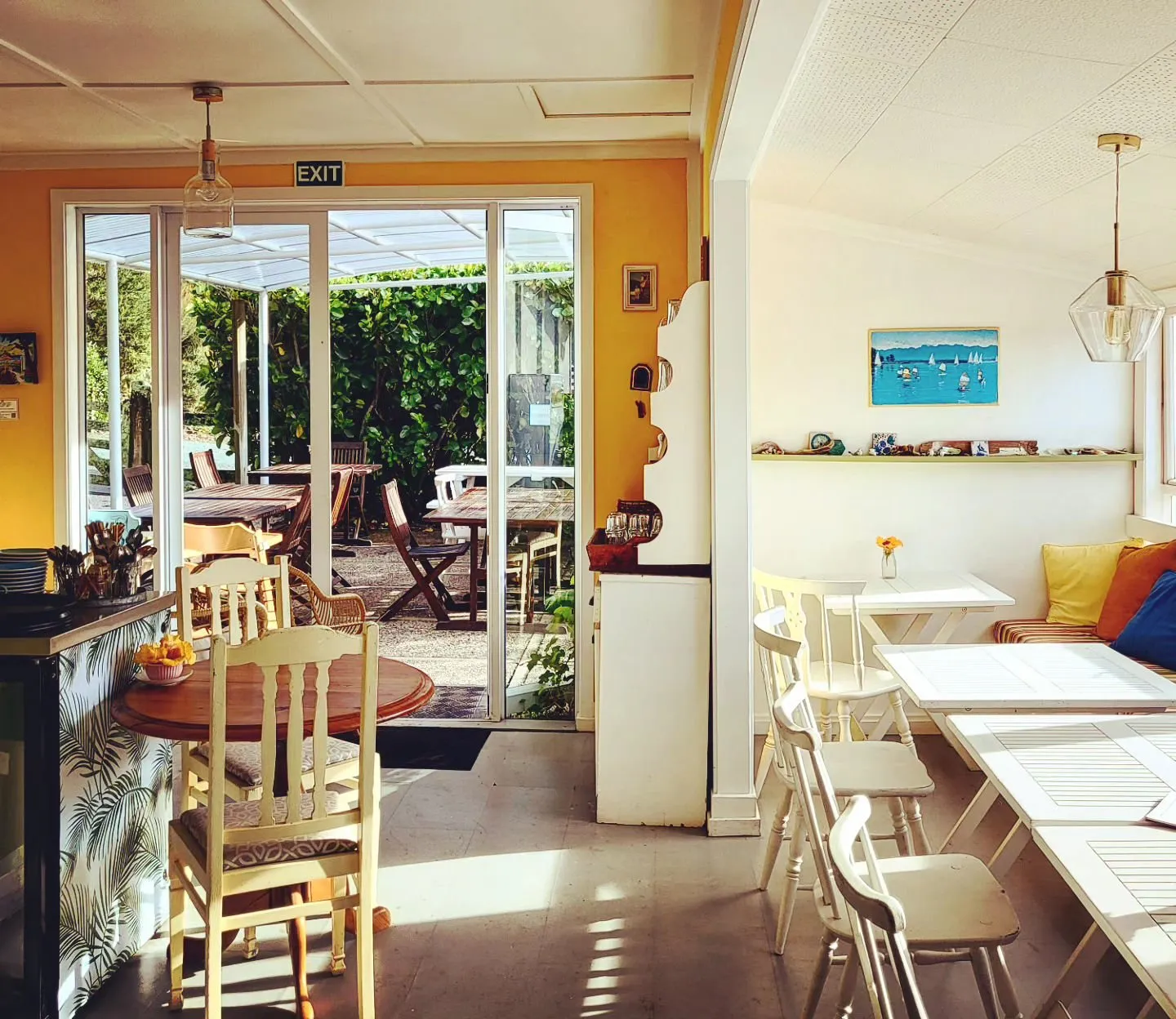 Sunlit café interior with white chairs and tables, yellow walls and open doors to the garden deck