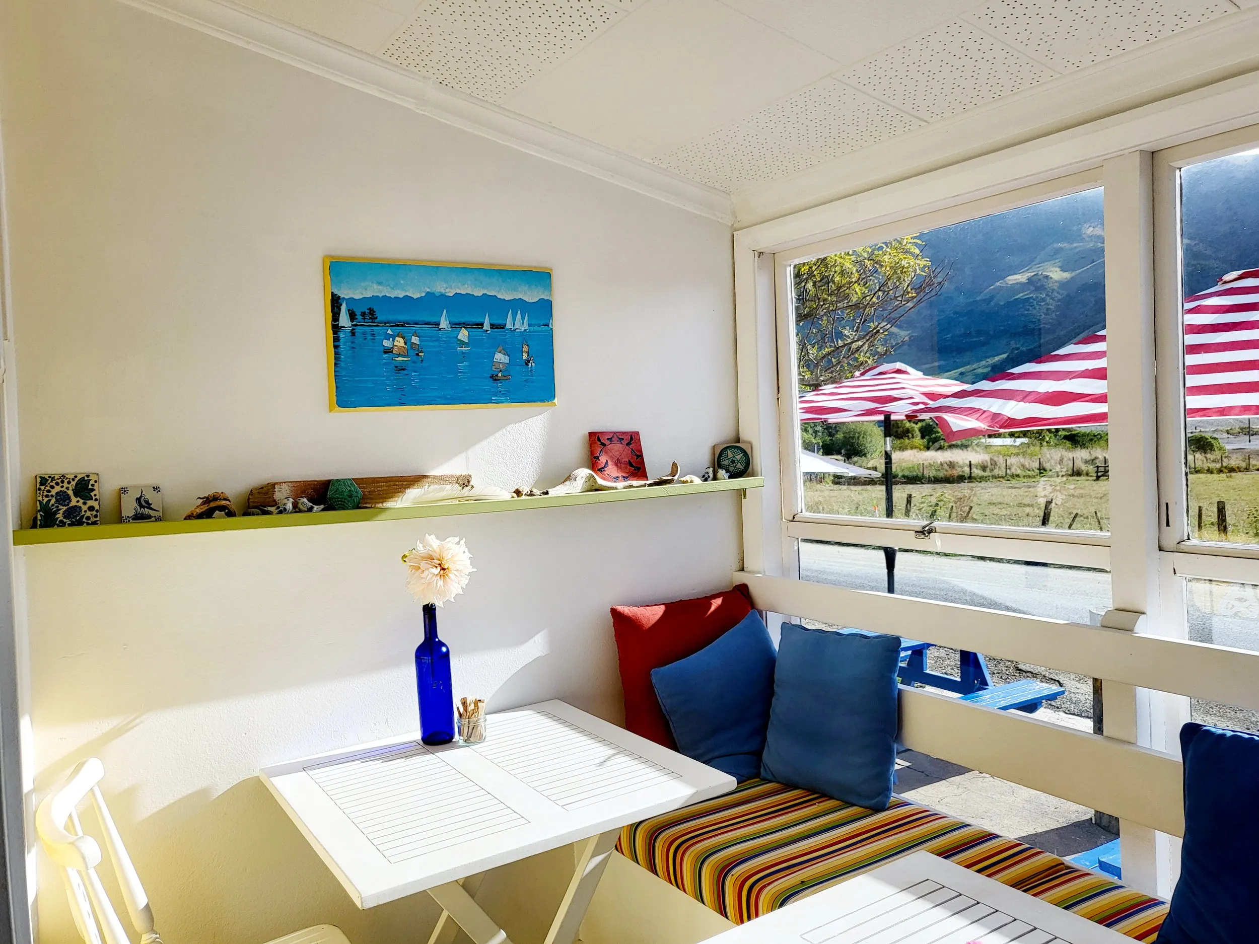 A window seat with blue and red cushions and a striped squab, looking out at striped umbrellas and green hills